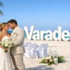 Alt text: Una coppia di sposi felici abbracciati su una spiaggia di Varadero, con sullo sfondo l’oceano e la scritta bianca “Varadero” in grande, tra decorazioni flore